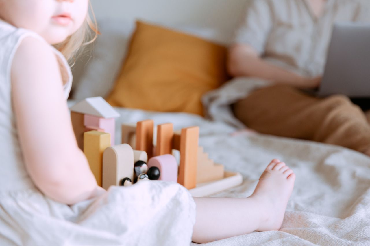 Crop faceless toddler girl in casual wear sitting on bed and playing with colorful wooden blocks while mother doing remote work with laptop on knees on background